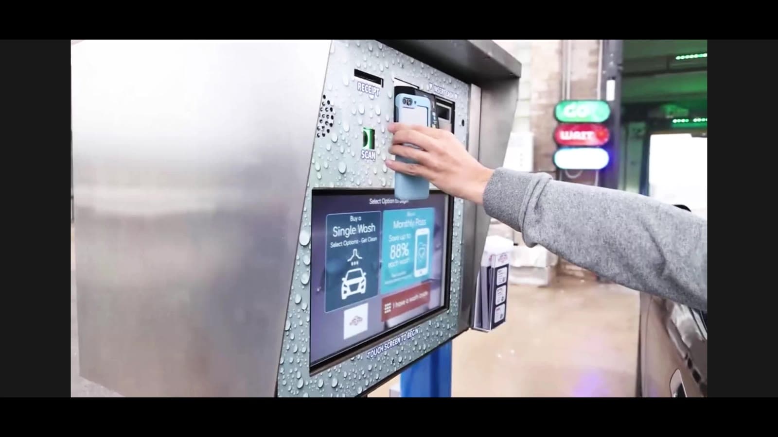 Customer holding a phone up to the scanner on a rain-covered car wash pay station showing single wash and monthly pass options